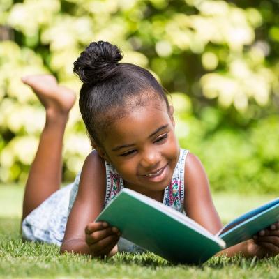 young child reading a book outside laying on the grass