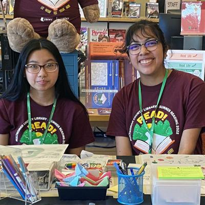 Two teen volunteers seated at the Summer Reading table inside the library