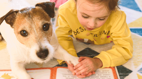 Photo of a child holding a dog's paw while reading to it from a book.