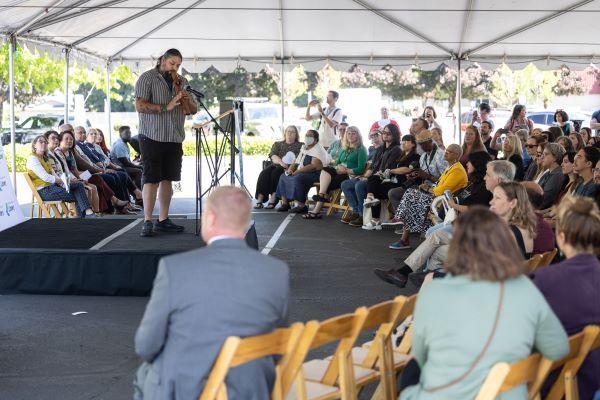 Man on a stage playing a wooden flute, audience of people sitting on chairs in a large tent.