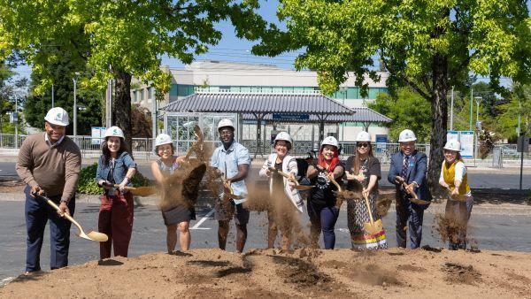 Nine smiling people standing behind a large pile of dirt, wearing hard hats, shoveling dirt to the air.