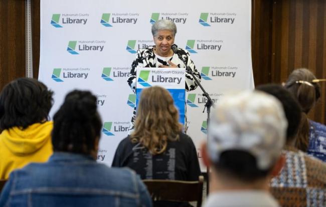 woman at a podium with an audience in front of her sitting in chairs.