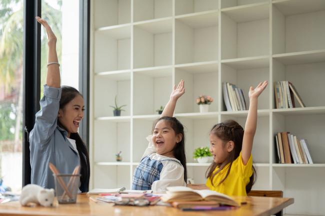 Adult with raised hand at table with books and two kids with raised hands.