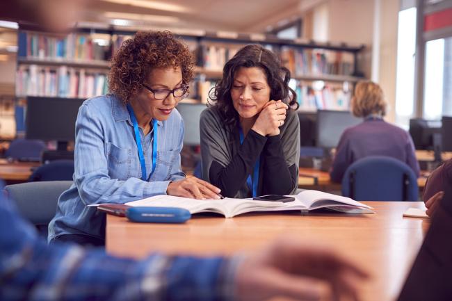 "Dos adultos mirando libros abiertos sobre una mesa en una biblioteca"