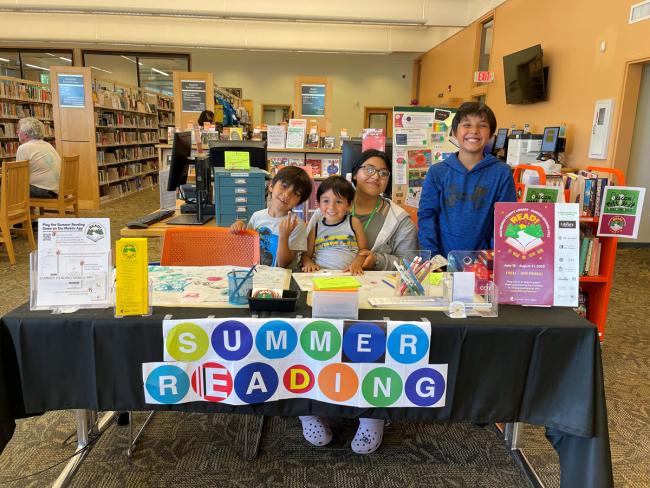 Voluntaria de Lectura de Verano con tres niños sentados detrás de una mesa con un cartel que dice Summer Reading (Lectura de Verano).