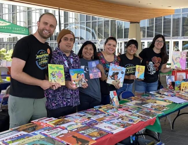 Seis miembros del personal de la biblioteca al aire libre, detrás de una mesa llena de libros, cada uno sosteniendo un libro y sonriendo.