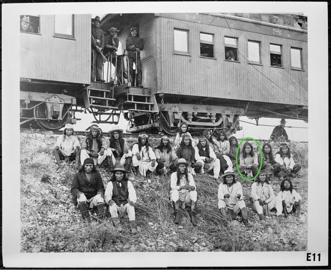 Historical photo of the Chiricahua Apache Prisoners, all sitting down outside in a group, with a train behind them.