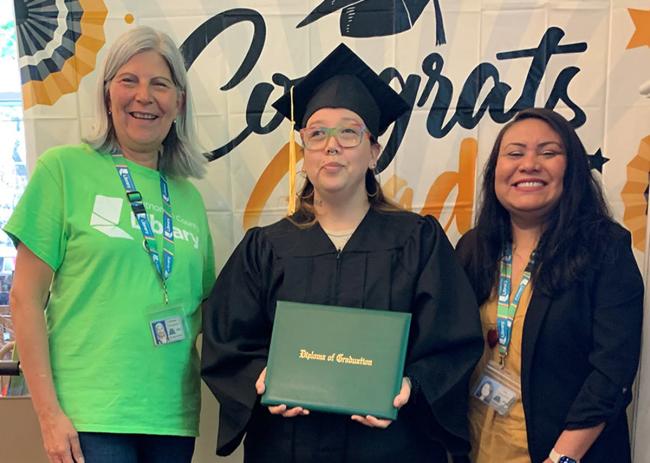 Three happy women stand together with the middle woman in graduation regalia and holding a diploma.