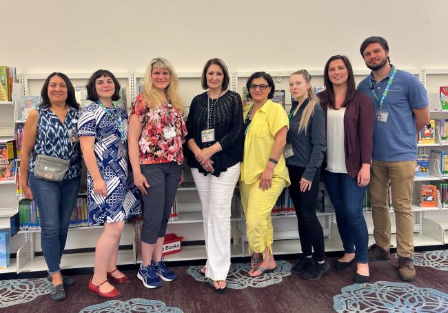 Eight people standing inside a library with a wall of bookshelves behind them. 