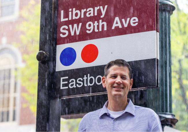 Man standing outside in the rain, behind him is a sign for the MAX train and Central Library.