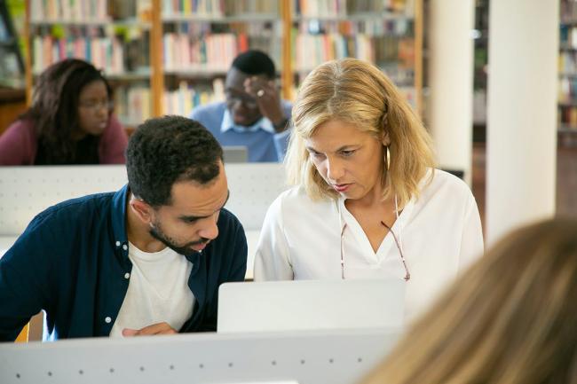 A woman on the right is looking at a computer with a man to her left inside a library.